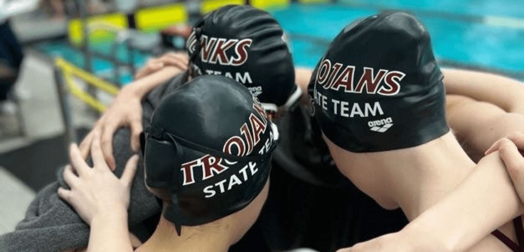 Three students with heads together, all wearing Jenks Trojan Swim Team swim caps.