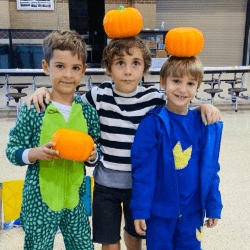 Three boys in costumes holding pumpkins in a cafeteria.