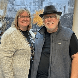 Anita Hardy, a white woman with short blonde hair standing next to Paul Hardy, a white man with white hair and a beard.