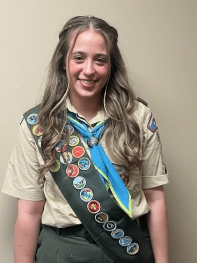 Kendall Ferguson, a teenage girl with long hair, wearing a Scouting USA uniform with neckerchief and merit badge sash.