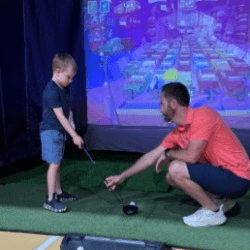 Derik Keim, a man with a beard and short dark hair, crouching down to instruct a young boy holding a golf club in front of a screen.