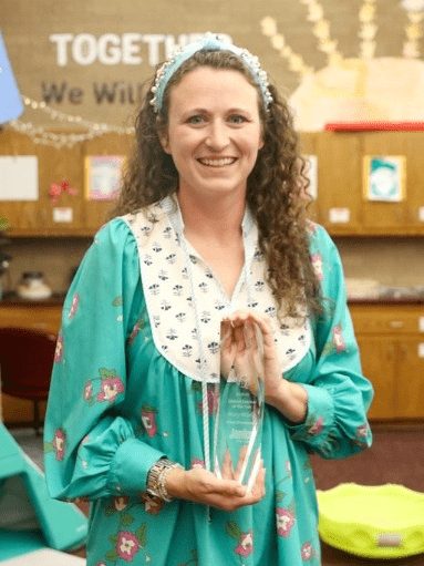 Macy Worley, a woman with long, curly brown hair, wearing a teal outfit, standing in her classroom holding a glass Teacher of the Year award.