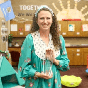 Macy Worley, a woman with long brown curly hair holding Teacher of the Year award standing in her classroom.