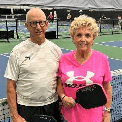 A white couple in athletic wear holding pickleball rackets on a court, Ed Cohlmia on left and Linda Cohlmia on right.